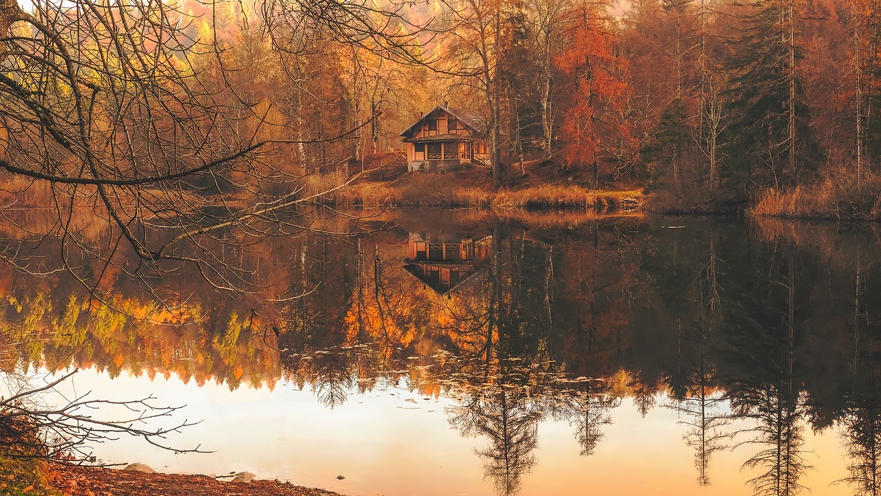 a home by a body of water in autumn surrounded by forest; colors seen are yellow, red, orange, and brown, as well as white from the reflecting water