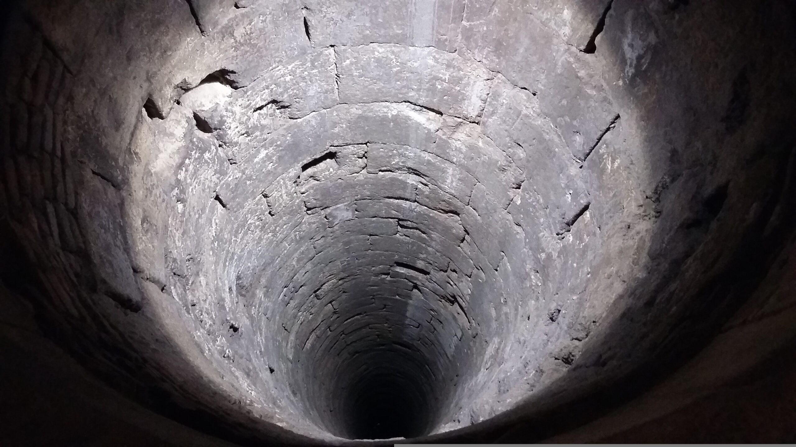 This image from Pixabay is used for the story "The Abyss," intended to represent the dark place the main character believes she is in. This image is of a round deep well or tunnel, in black and white, with cement walls and an undiscernible black bottom to the structure. 